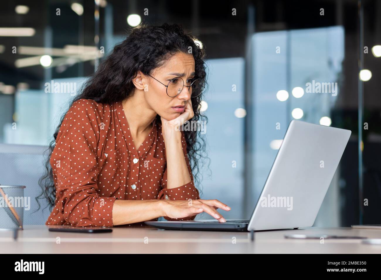 Tired and upset young Hispanic woman sitting in office at desk in front ...