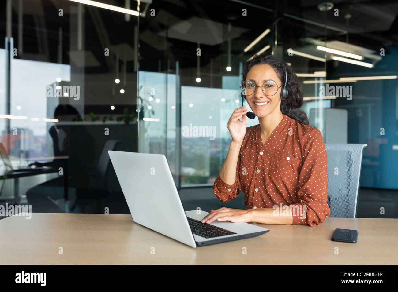 Portrait of a young beautiful Latin American woman teacher. Sitting at a desk in a headset. Teaches online, distance learning through a video call from a laptop. He looks at the camera with a smile. Stock Photo