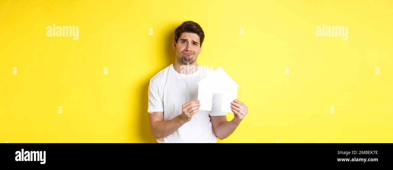 Real estate concept. Displeased young man showing paper house model and grimacing upset, standing over yellow background Stock Photo