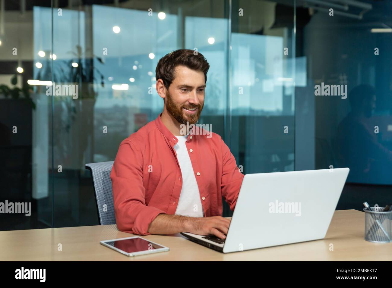 Successful businessman in red shirt happily working with laptop inside office, mature man with beard at workplace typing on keyboard smiling satisfied with work results and achievement. Stock Photo