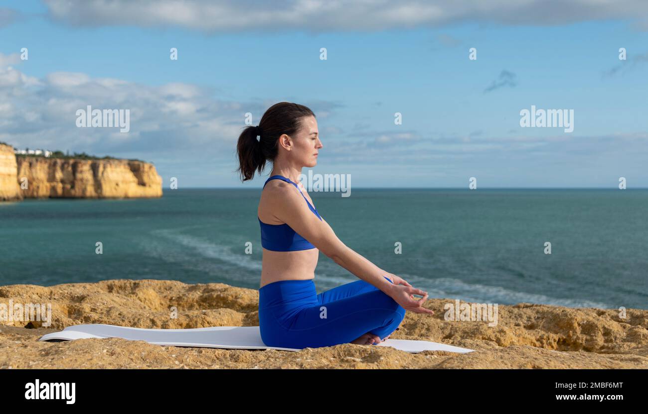 Fit woman meditating by the ocean, sitting on a yoga mat. Stock Photo