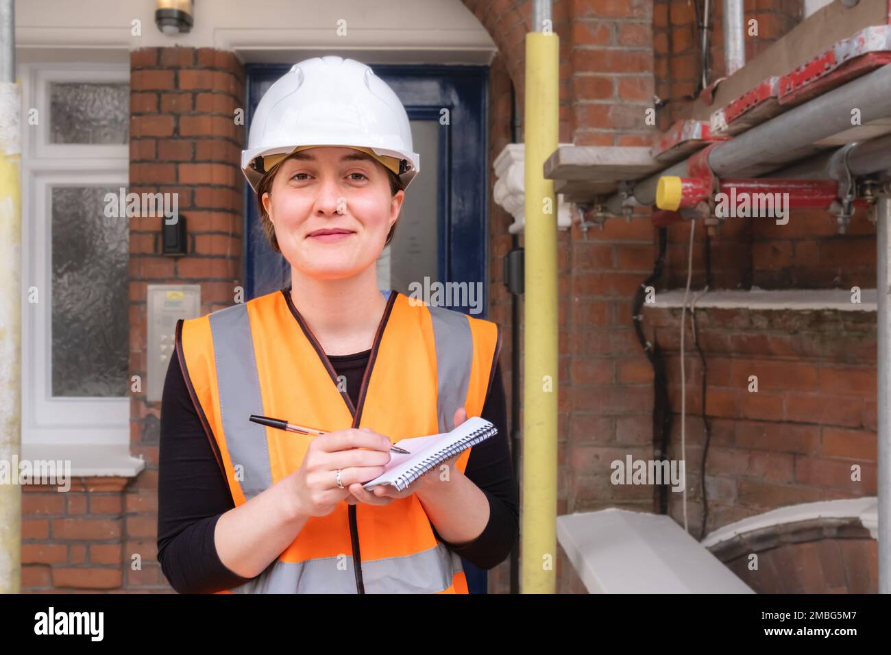 A civil engineer lady inspecting and supervising a construction site building with scaffolding, wearing a hard hat and orange personal protective equi Stock Photo