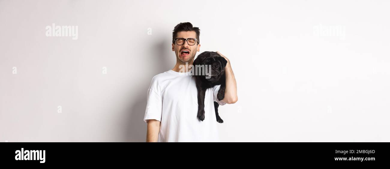 Sad dog owner crying, holding cute black pug on shoulder and looking miserable, sobbing while standing over white background Stock Photo