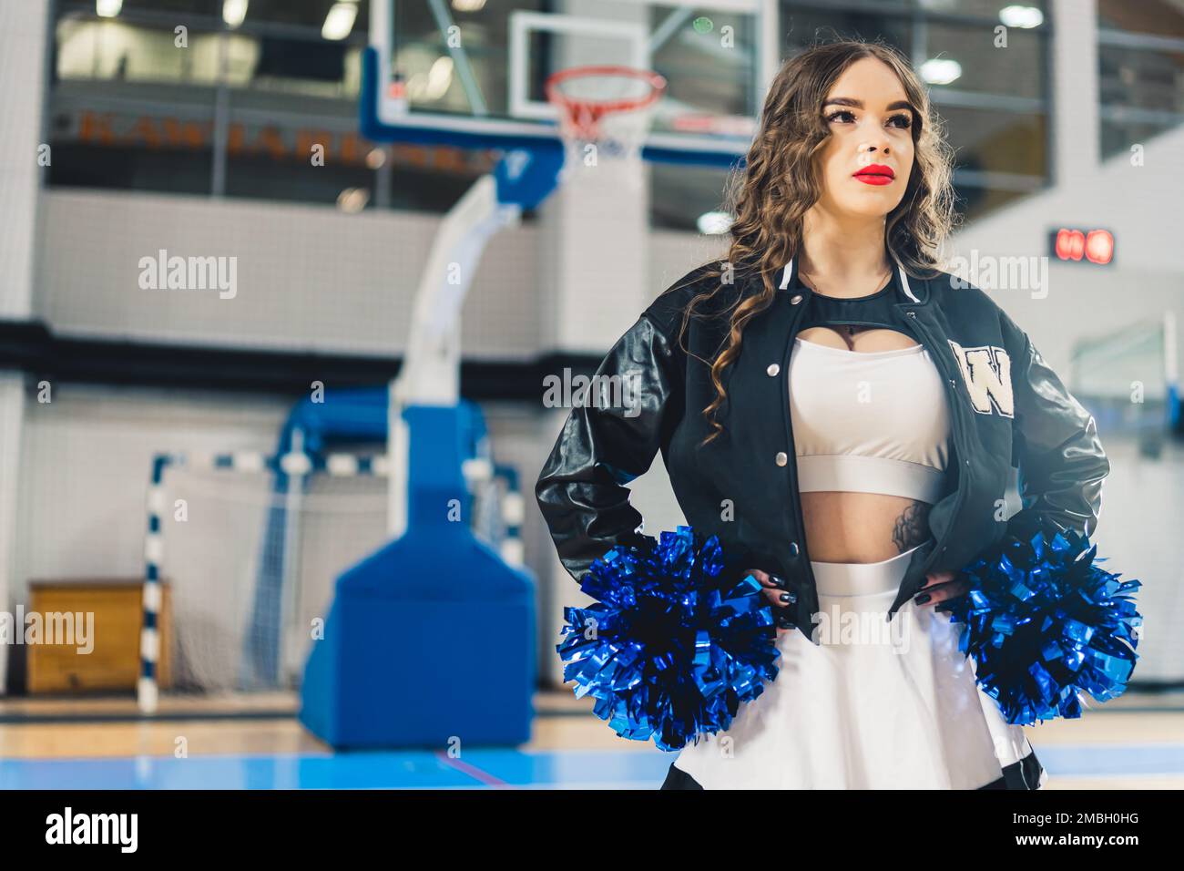 Horizontal shot of cheerleader wearing red lipstick in a jacket posing with blue shiny pom-poms. Basketball court blurred in the background. High quality photo Stock Photo