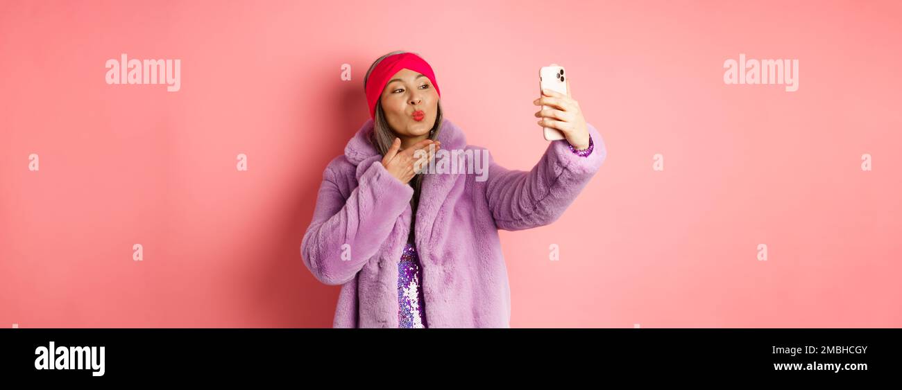Fashion concept. Beautiful and trendy asian mature woman in faux fur coat with party dress, taking selfie, sending kiss to smartphone camera, posing Stock Photo