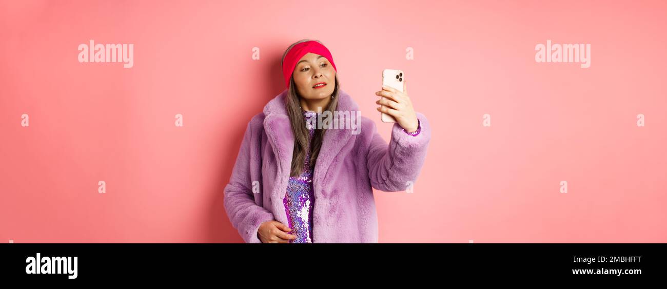 Fashion concept. Stylish asian senior female taking selfie on smartphone, posing in purple faux fur coat and party dress, standing over pink Stock Photo