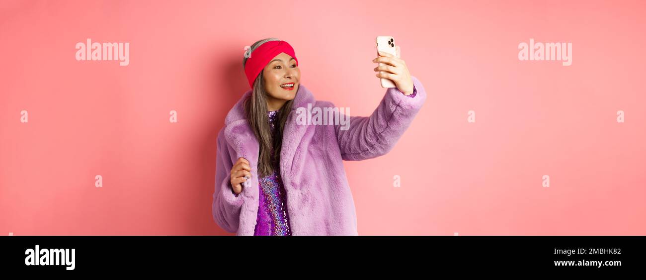 Fashion concept. Beautiful asian senior lady taking selfie on smartphone, posing in party glittering dress and purple faux coat, standing over pink Stock Photo