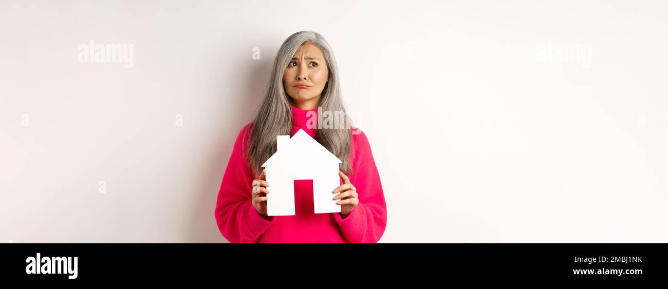 Real estate. Sad asian senior woman dreaming about own property, holding paper house model and grimacing upset, looking at upper left corner, white Stock Photo