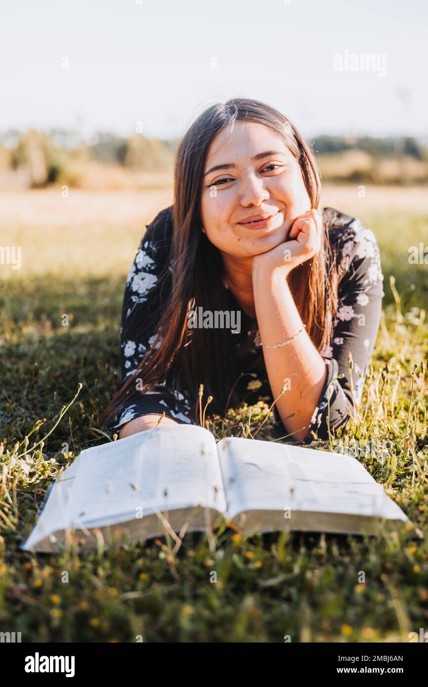 Smiling christian girl lying on the grass reading her bible, in the field at sunset. Looking at
