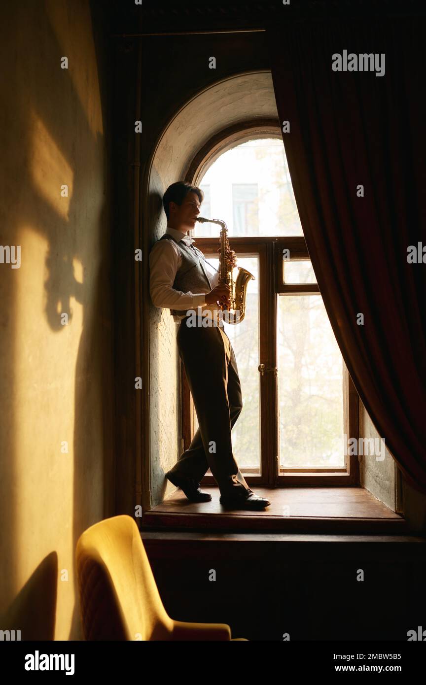 Creative young man playing saxophone standing on windowsill Stock Photo