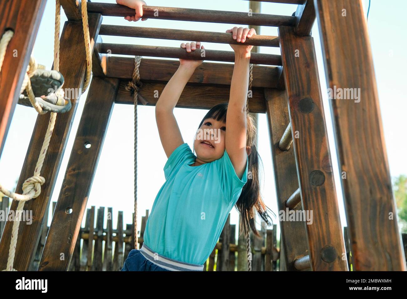 Little girl playing on playground, hanging on a horizontal bar. Active little girl exercising outdoors, hanging on monkey bars in the playground. Stock Photo