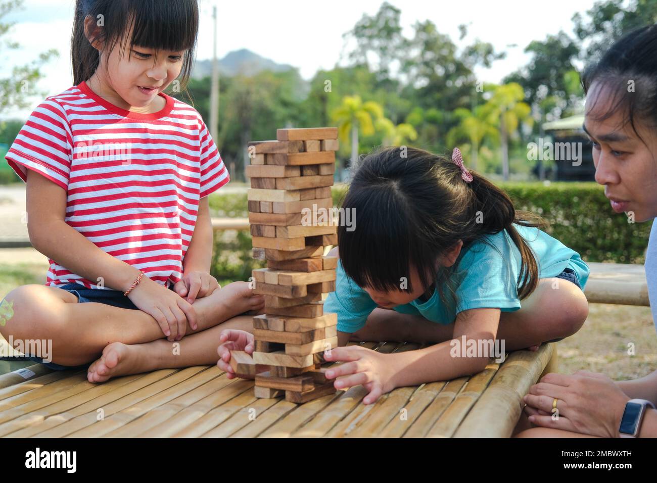Excited kids and mom playing Jenga tower wooden block game together in the park. Happy family with children enjoying weekend activities together. Stock Photo