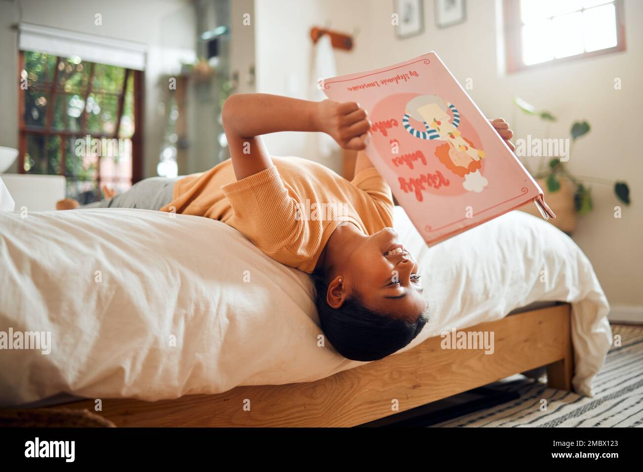 Happy child reading book on bed for home learning, language education and creative development upside down. Smart, intelligent and Indian kid with Stock Photo