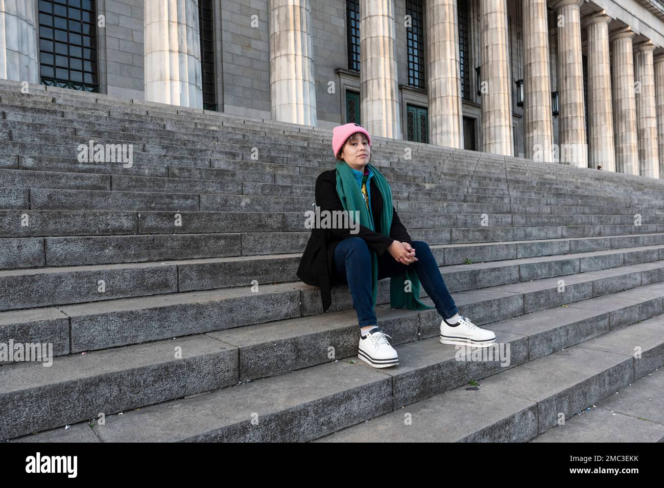 Female latin student with brown hair and pink hat sitting on the stairs ...