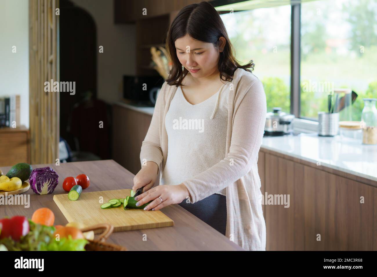 Pregnant Asian woman cutting cucumber for fresh green salad, female prepares tasty organic dinner at home, healthy nutrition for future mother Stock Photo