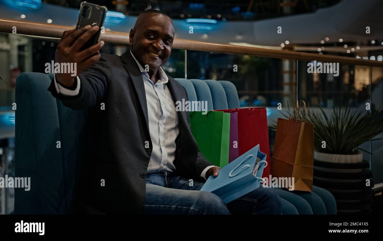 Middle-aged male African American man buyer client customer blogger influencer vlogger with shopping bags in mall video call make photography with Stock Photo