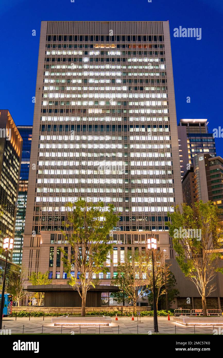 The Marunouchi Centre Building seen from Marunouchi Ekimae Square at night time. Tall imposing building illuminated with several trees in front. Stock Photo