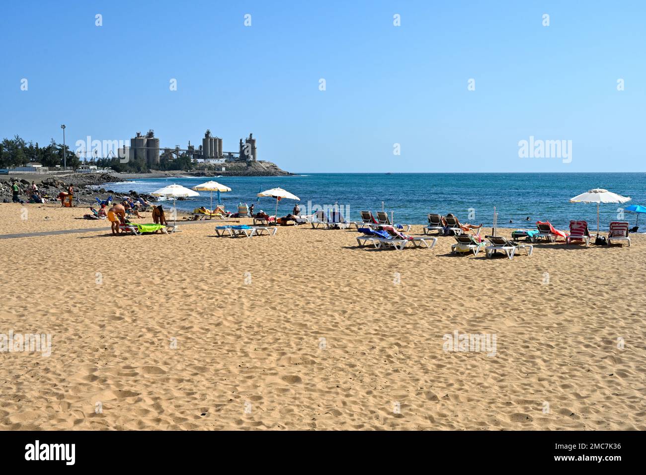 Sandy beach with tourists, Playa Las Marañuelas, town of  Arguineguín with cement factory in distance, Gran Canaria, Canary Islands Stock Photo