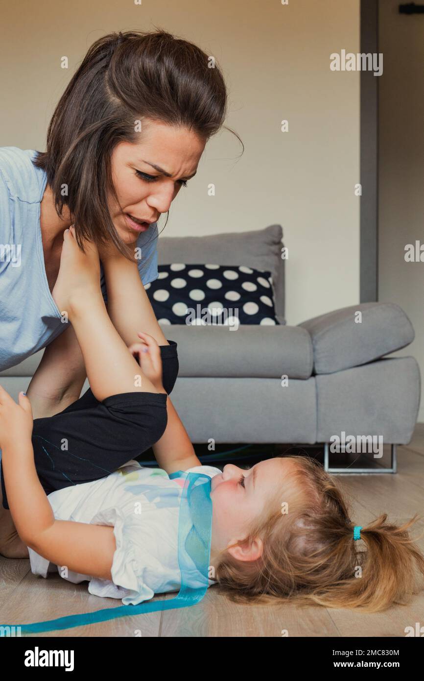 as they play tickle game in the living room, the little girl puts her feet on her mom's chest, who pretends to feel sick to make her laugh more Stock Photo
