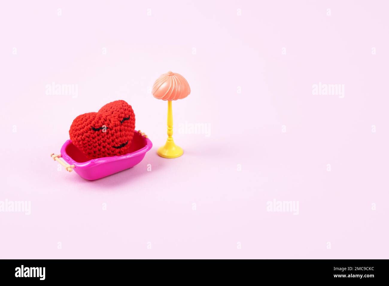 Concept of Valentine's Day. Knitted red heart in a pink toy bath and a toy lamp on a lilac background. Symbol of love, family, loyalty Stock Photo