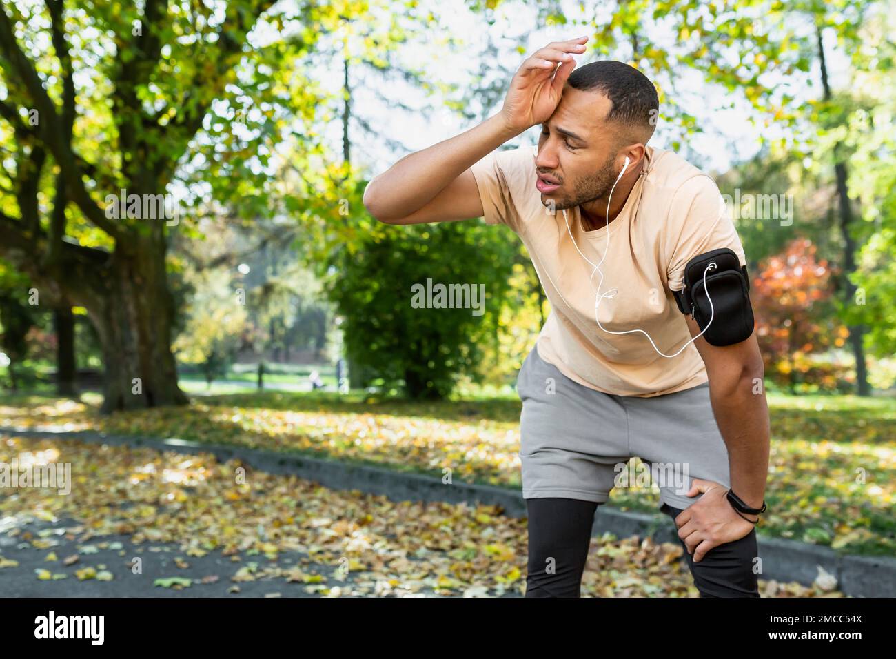 Tired man resting in park after jogging and fitness class, hispanic man using phone listening to music and podcasts in headphones breathing tired and exhausted. Stock Photo