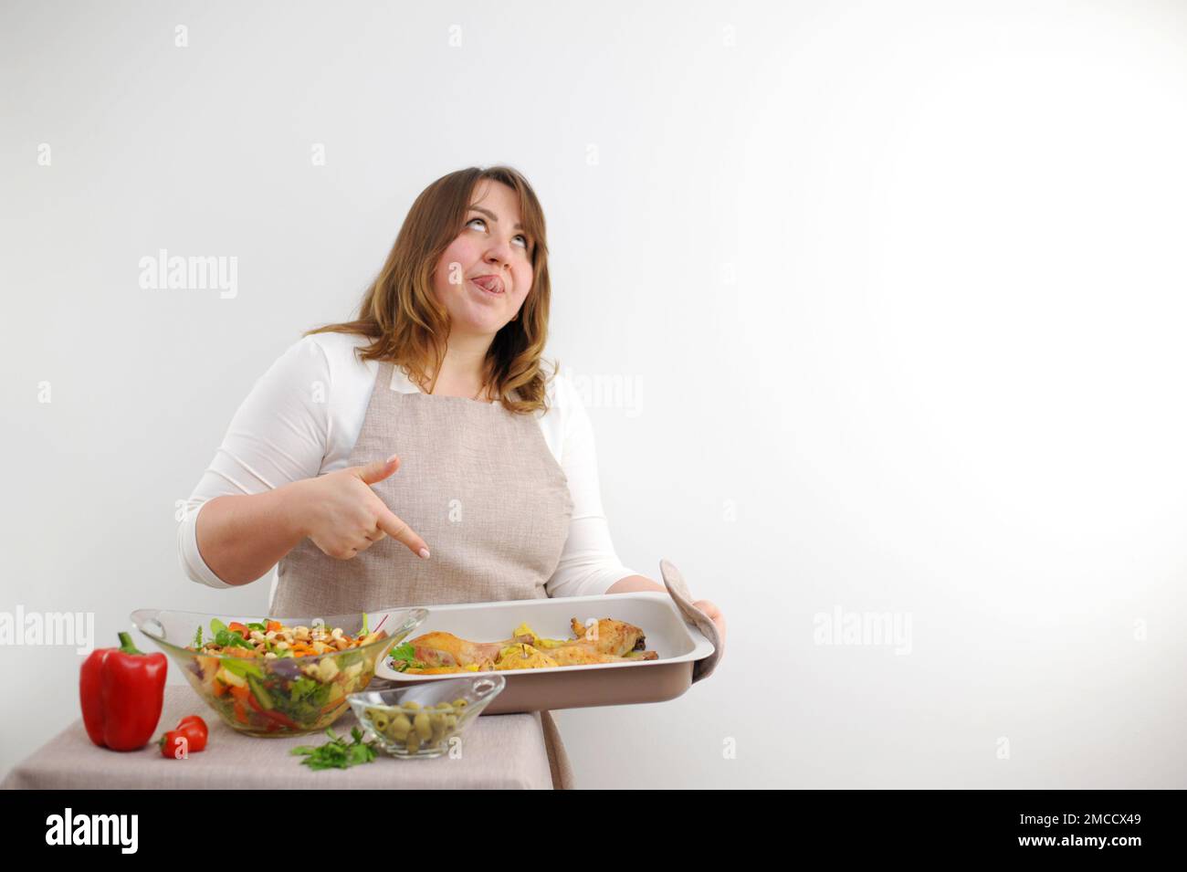 funny facial expression at cook puffy woman rolled eyes stuck tongue out licks lips with pleasure points to baking sheet in which dinner is cooked lunch brightly colorful salad with nuts ingredients Stock Photo