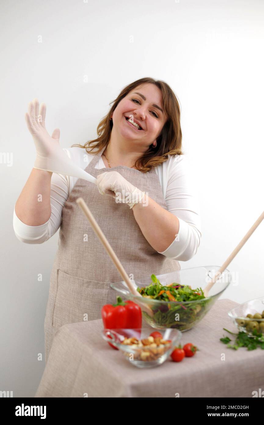 beautiful plump woman She cooks food in gloves wearing gloves looks into frame smiles concept of cleanliness neatness healthy attitude to cooking restaurant food on white background beige apron Stock Photo