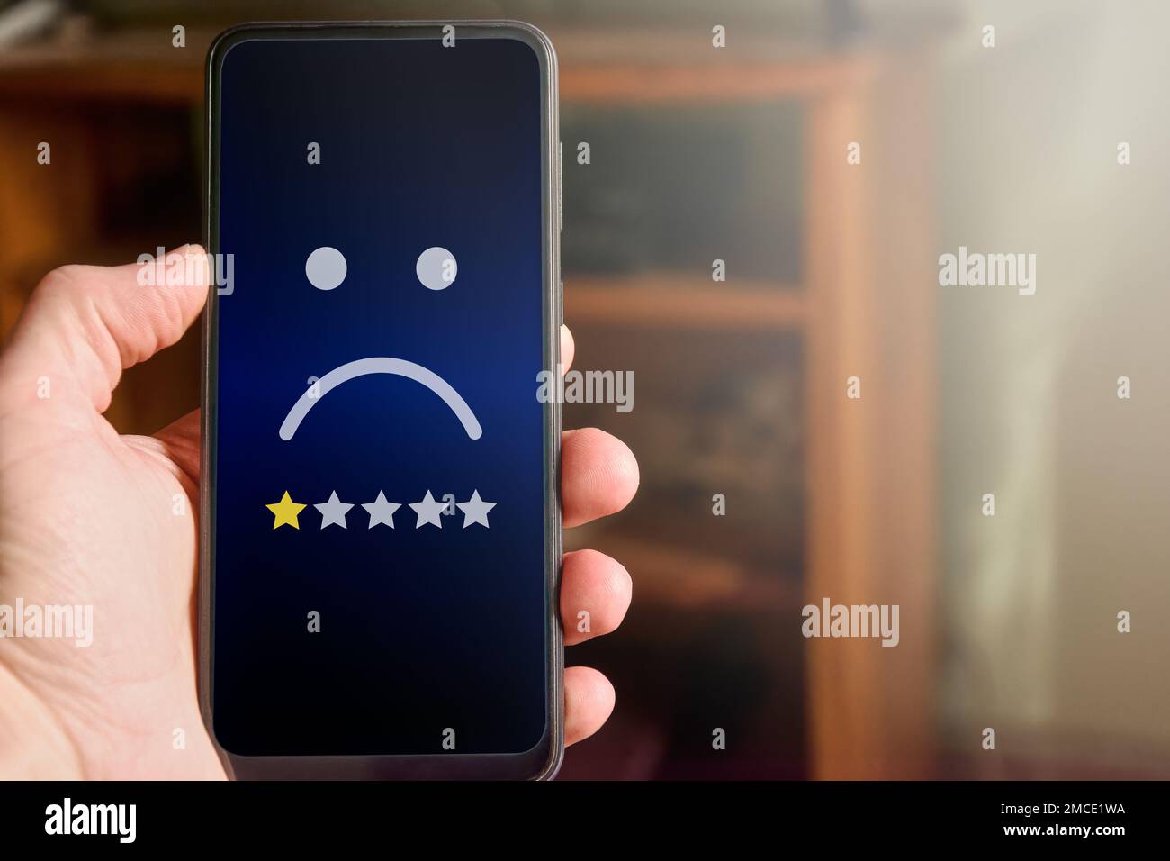 Closeup of male hand holding mobile phone with sad face and one star rating. Very unhappy customer giving negative feedback. Customer experience, clie Stock Photo
