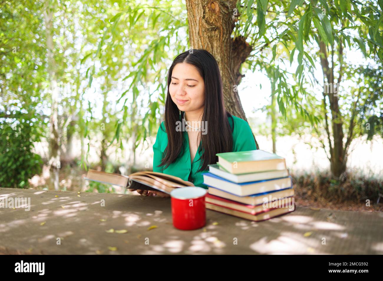 Cute young woman reading books in the field under a willow tree, with a coffee mug. World book day Stock Photo