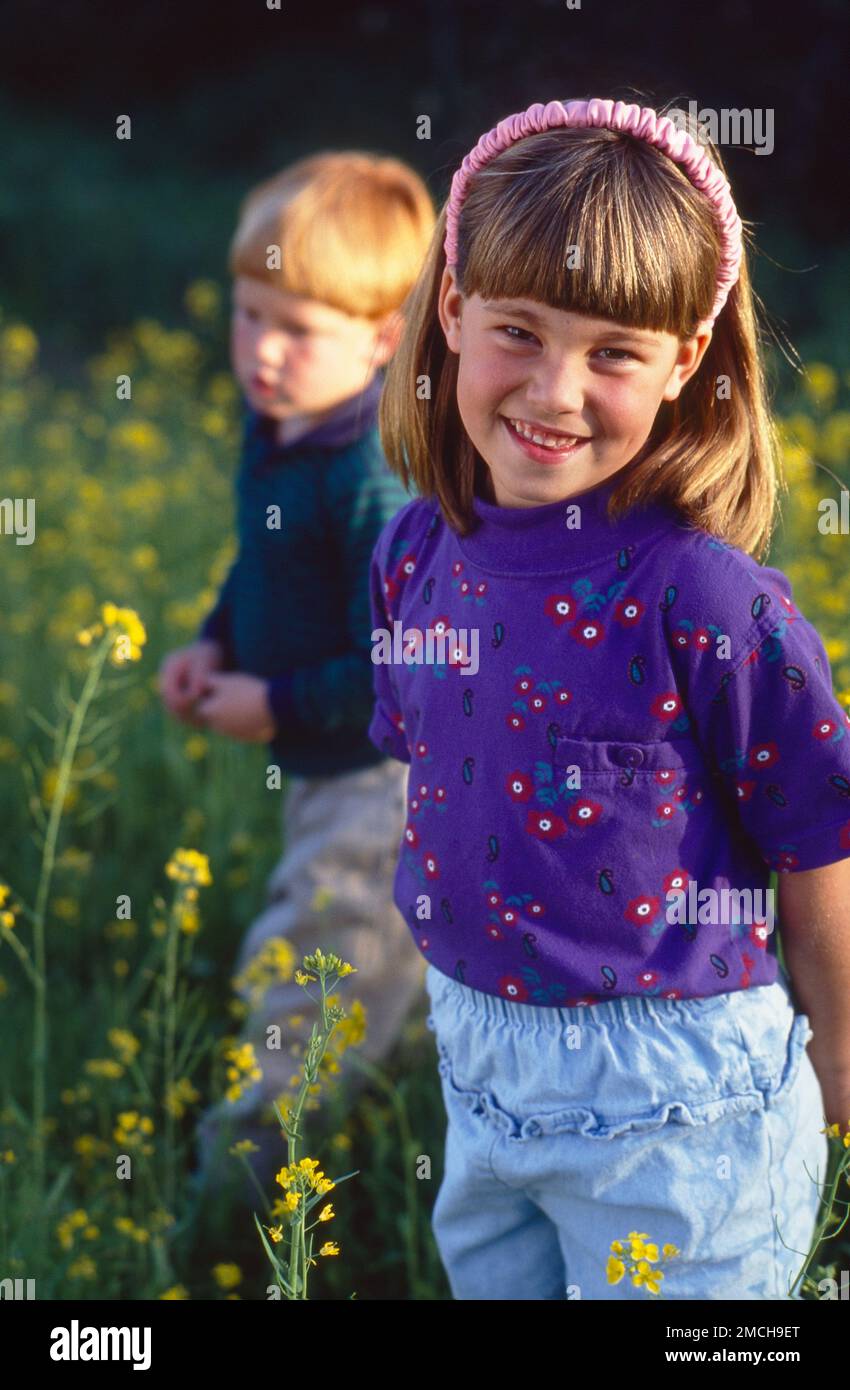 Little redheaded boy and girl standing out in the field of tall grass and yellow flowers grow. Looking at the camera boy in the background Stock Photo