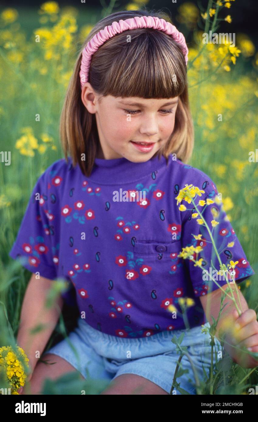 Young girl child's outdoors, sitting in a field of tall grass with yellow flowers, glancing down looking at the flower Stock Photo
