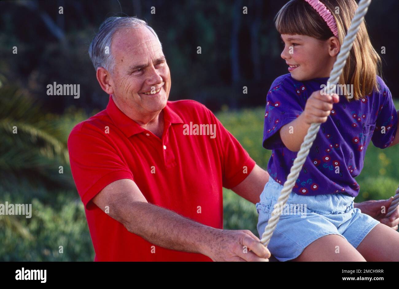 Grandpop pushing girl, grandchild on a swing both looking at each other smiling Stock Photo