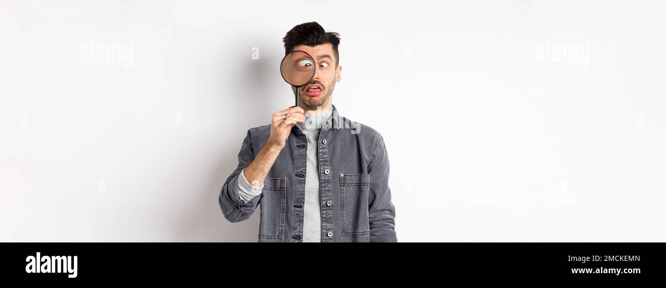 Funny young man squinting, showing faces with magnifying glass, standing on white background Stock Photo