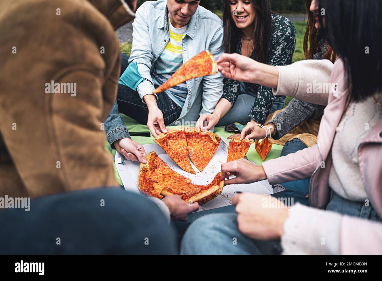 A group of friends sit on the grass and enjoy a large pizza together. The focus of the shot is a close-up of the pizza slices being held by the indivi Stock Photo