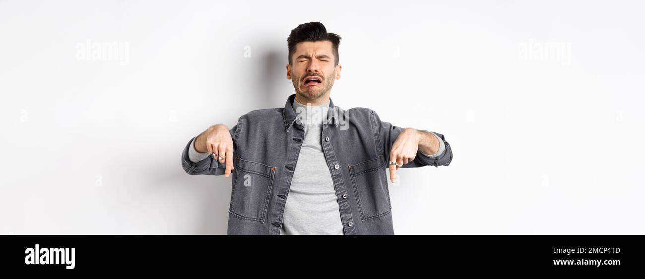 Sad crying man pointing fingers down, sobbing miserable, showing upsetting news, standing in denim jacket on white background Stock Photo