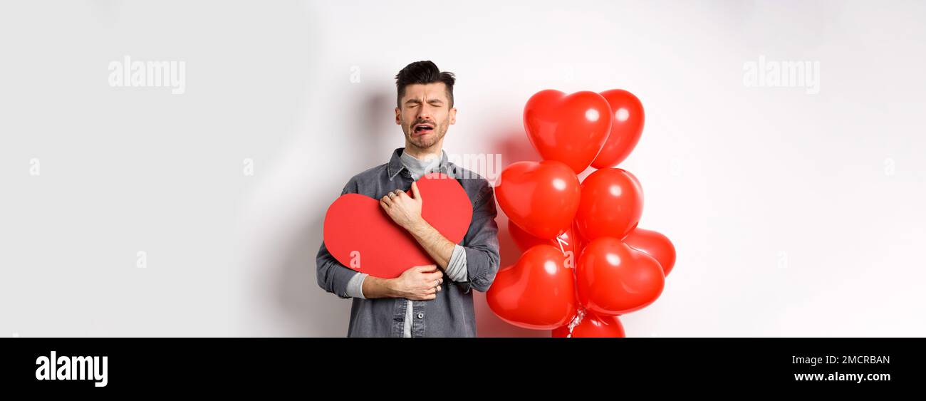 Crying man standing single and lonely on Valentines day, hugging heart cutout and sobbing miserable, being heartbroken and rejected by lover, white Stock Photo