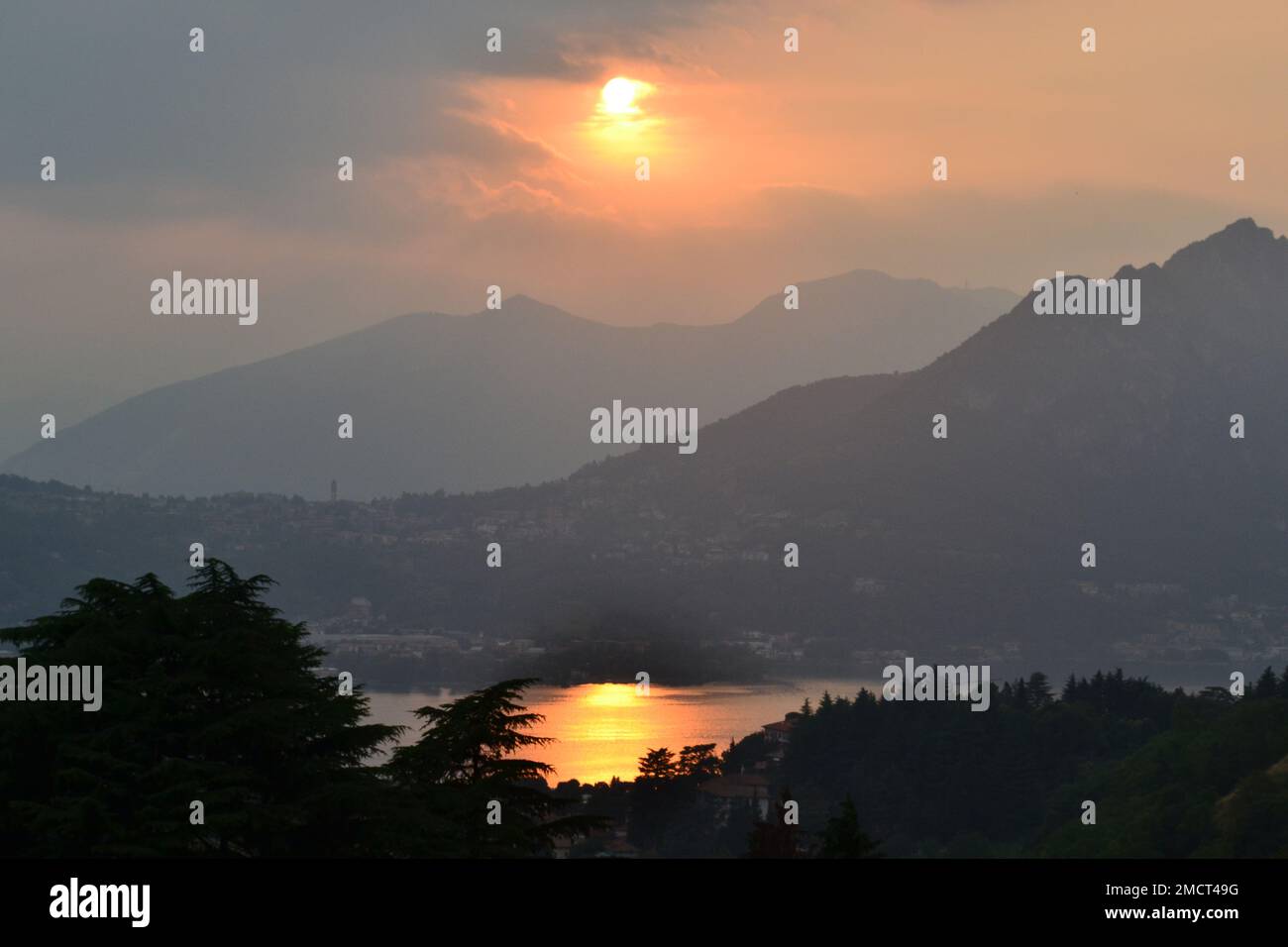 Panoramic view of the beautiful sunset over lake and mountains. Sunset sun rays coming out of the cloudy sky towards multi silhouette mountain ranges. Stock Photo