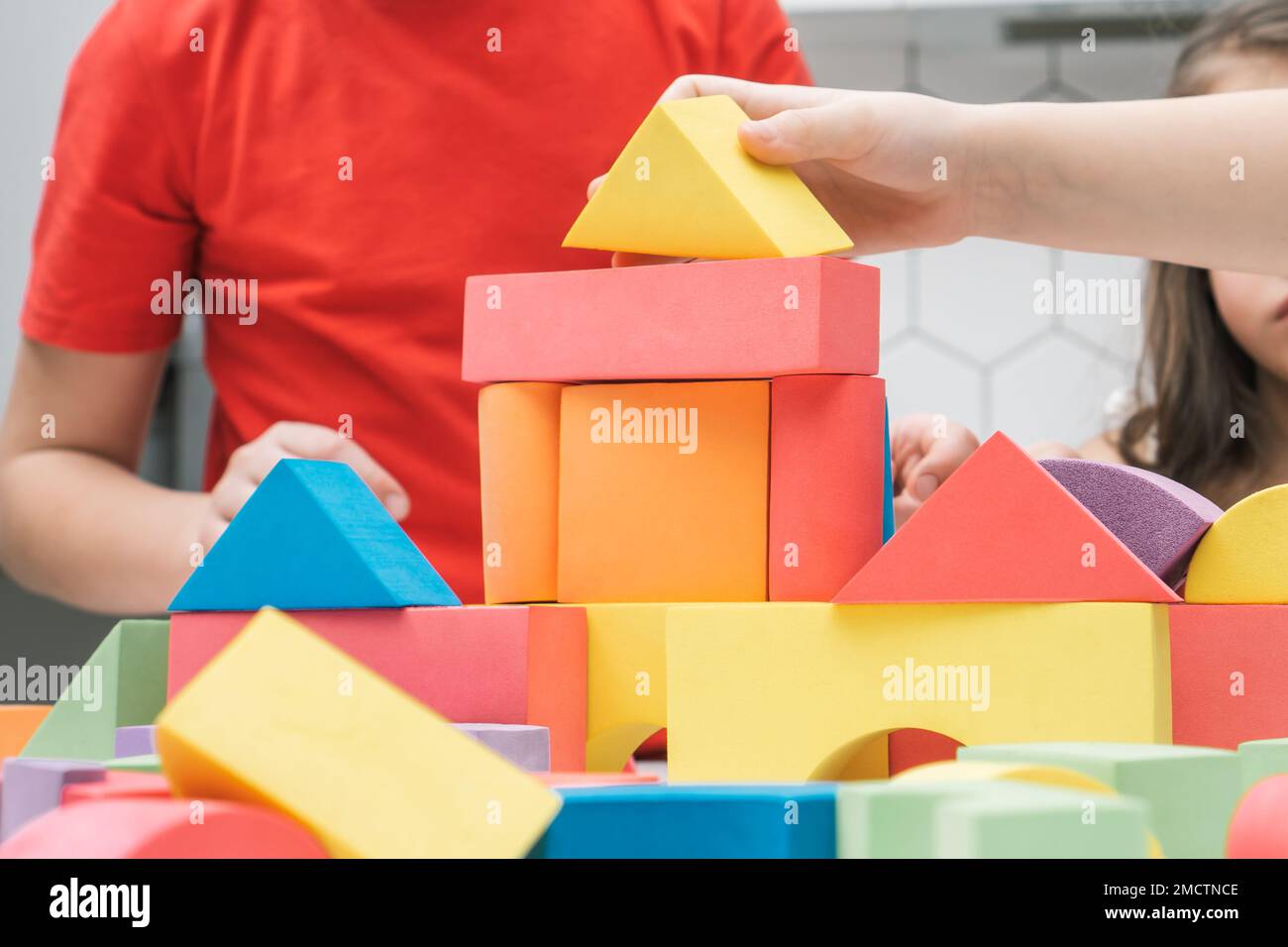 Cropped photo of preteen children friends sitting at table in kitchen, building tower house castle of colorful wooden construction blocks geometric fi Stock Photo