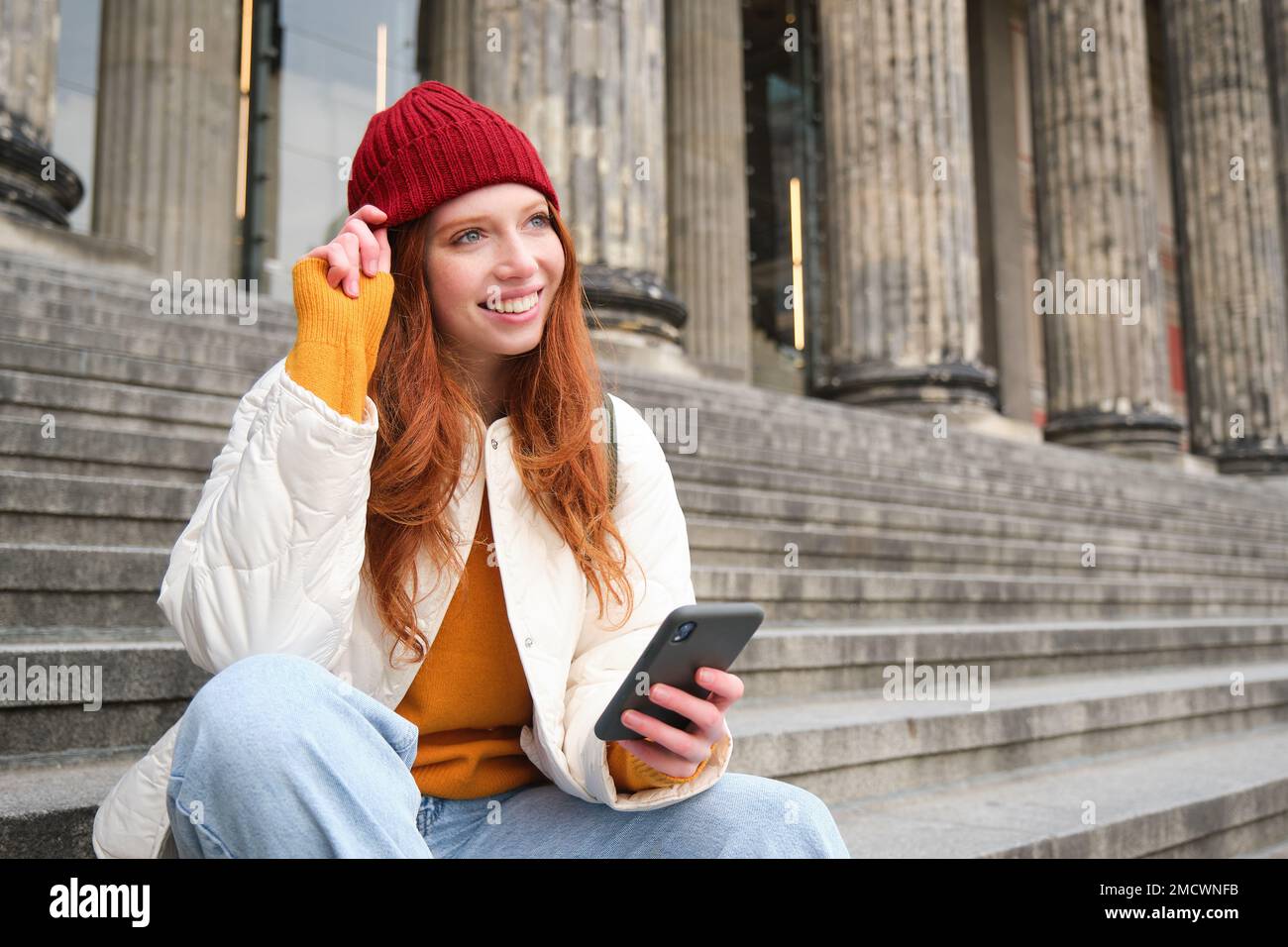Stylish young redhead woman, talking on mobile phone app, using social media application, looking for something online on smartphone, sits on stairs Stock Photo