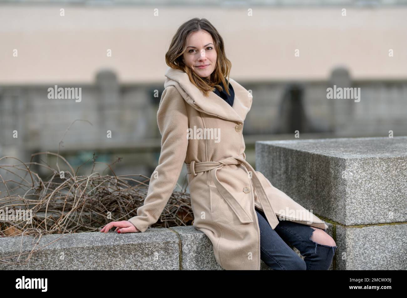 Pretty young woman with long brunette hair, a light brown cloth coat and torn black trousers sits on a wall of natural stones Stock Photo