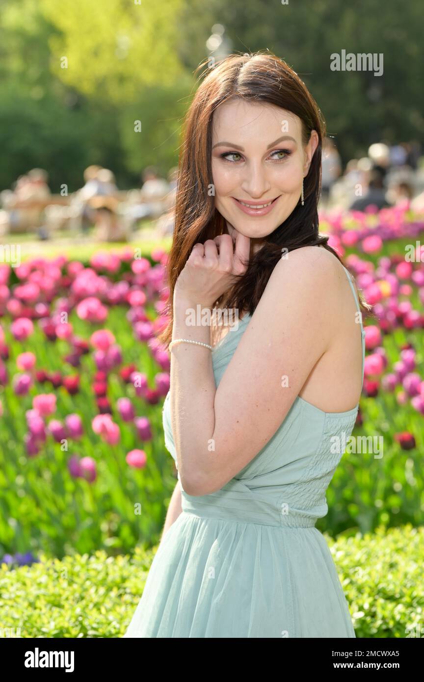 Standing, smiling pretty young woman in an off-the-shoulder turquoise dress in a spring-like park with blooming flowers. The expectant gaze with open Stock Photo