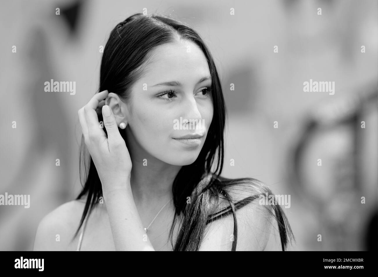 Black and white portrait of a very young pretty woman with a blurred background. With her right hand she guides her long hair behind her ear Stock Photo