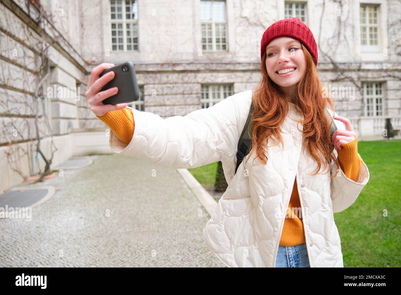 Portrait of happy girl tourist, takes selfie on smartphone in front of historical building, posing for photo on mobile phone camera Stock Photo
