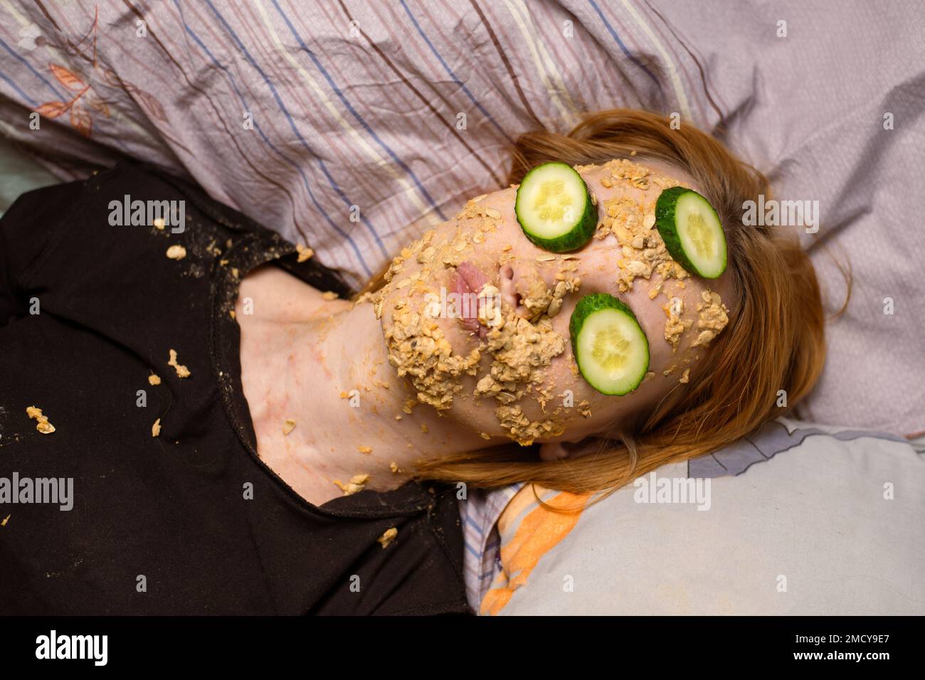 A woman makes a cosmetic mask at home from oatmeal on her face and cucumber. Anti-aging treatments, skin care concept. Stock Photo
