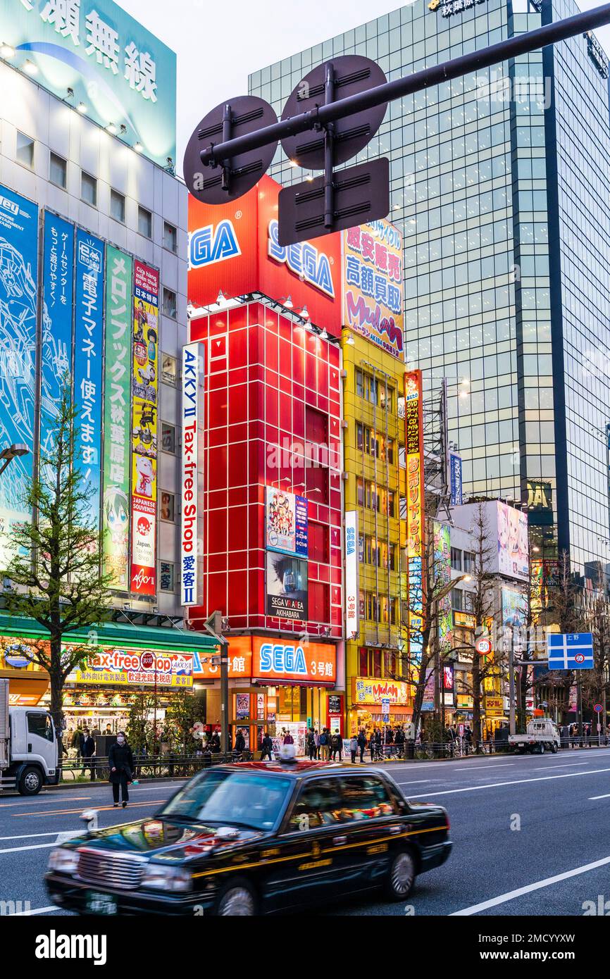 View along Chuo Dori, the main shopping street in Akihabara, Tokyo. Buildings illuminated, taxi in the foreground and the Sega Red building behind. Stock Photo