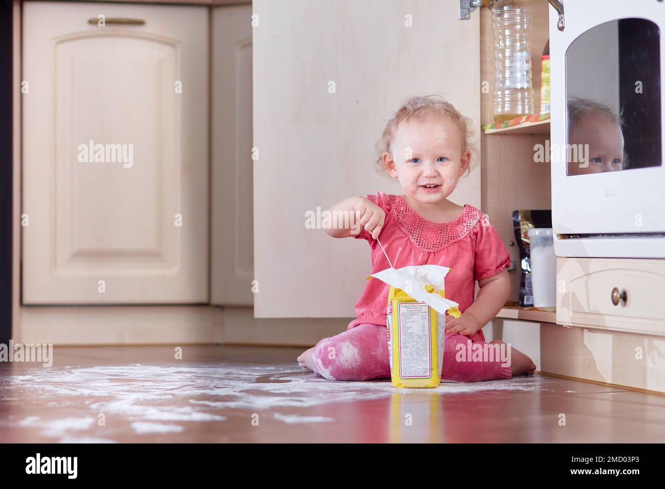 A child sits in a dirty heap of flour scattered on the floor in the kitchen at home. Copy space Stock Photo