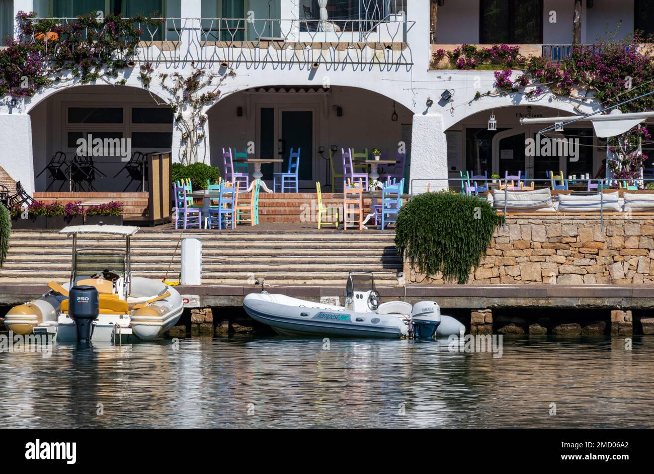 Marina dell' Orso di Poltu Quatu Waterfront. Hotel and Restaurant with Bright Coloured Chairs, Moored Dinghies and Reflections. Sardinia, Italy. Stock Photo