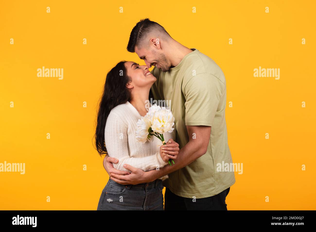 Happy millennial arabic guy hugging lady, gives bouquet of flowers, isolated on orange background, studio Stock Photo