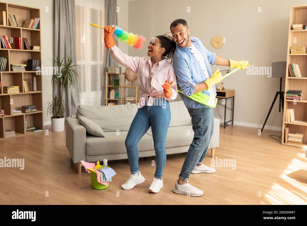 Happy joyful black husband and wife tidying home and singing during housecleaning, using mop and duster as microphones Stock Photo