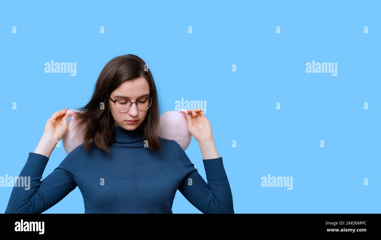 Portrait of a student girl in glasses and a sweater sleeping in a travel pillow on a blue background, banner, copy space. A woman at rest with a pillo Stock Photo